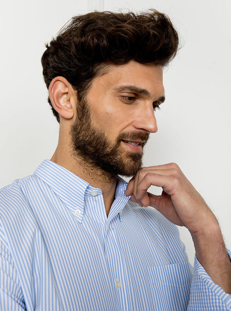 Man wearing long sleeved shirt with sky blue and white thin stripe and the inside collar and cuffs trimmed with gingham pattern. Made with 100% cotton in Thomas Mason fabric.
